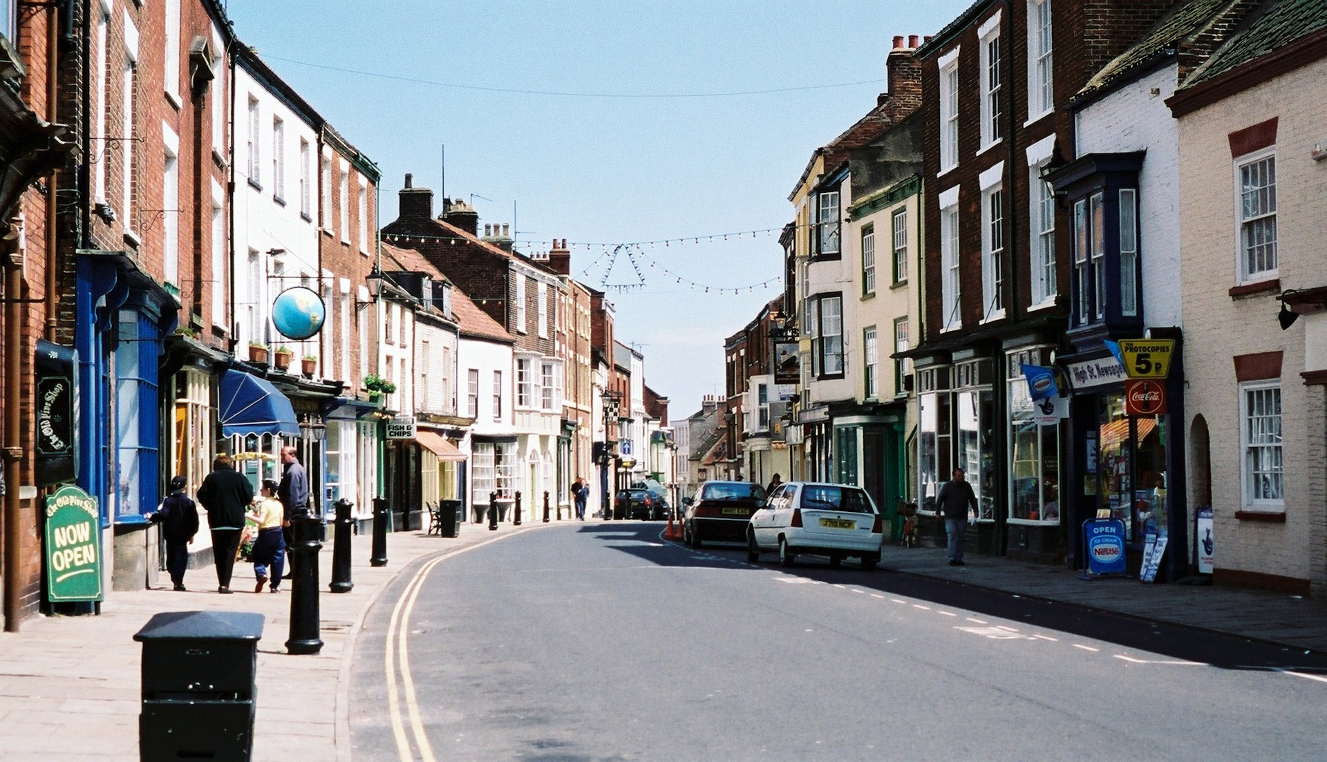 A photograph of a British high street in 1999 showing shops, cars, and pedestrians.