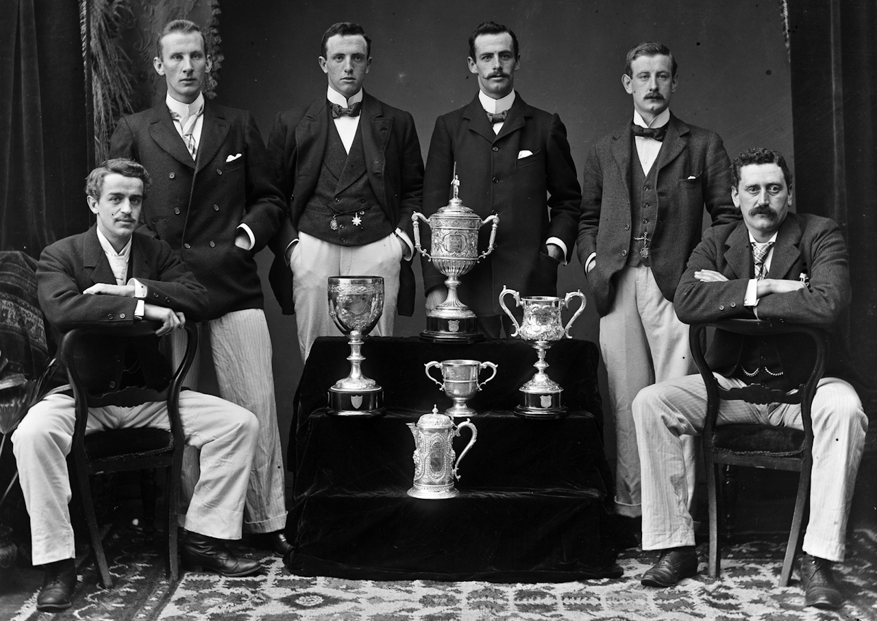 19th Century photo showing six men standing and sitting next to several trophies.