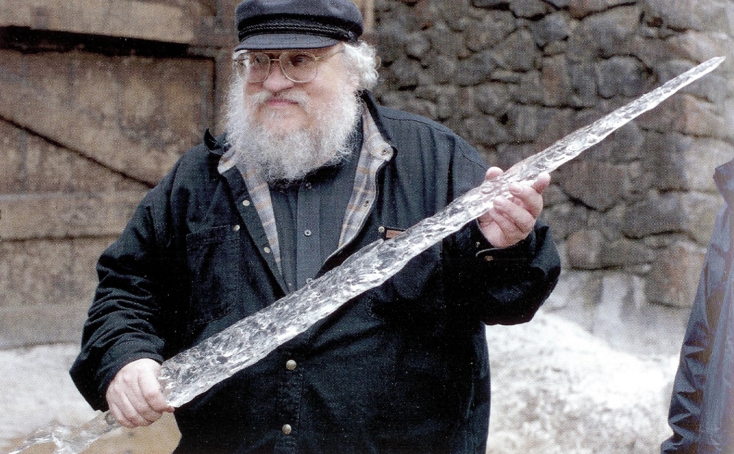 Photo of George R R Martin holding a large ice sword prop on the set of Game of Thrones.