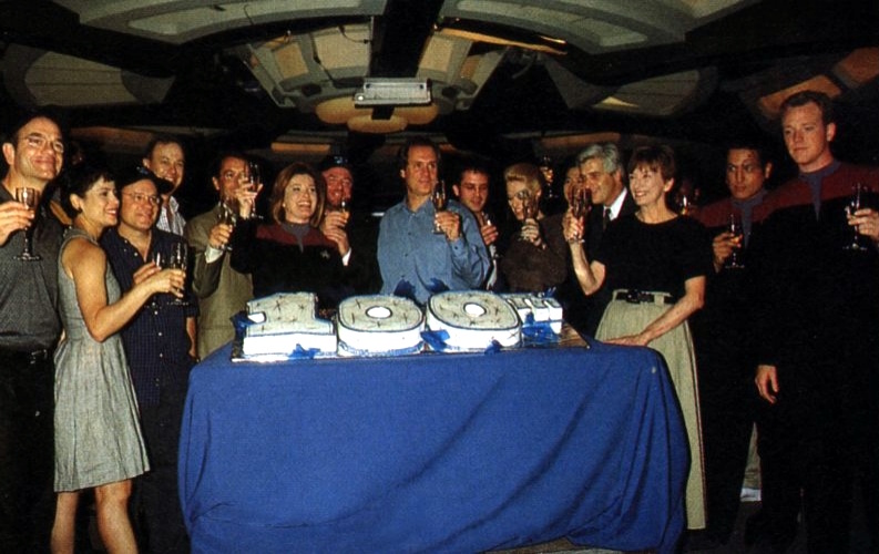 Behind-the-scenes photo from Star Trek: Voyager's 100th episode celebration. A large cake is present along with the cast and crew.