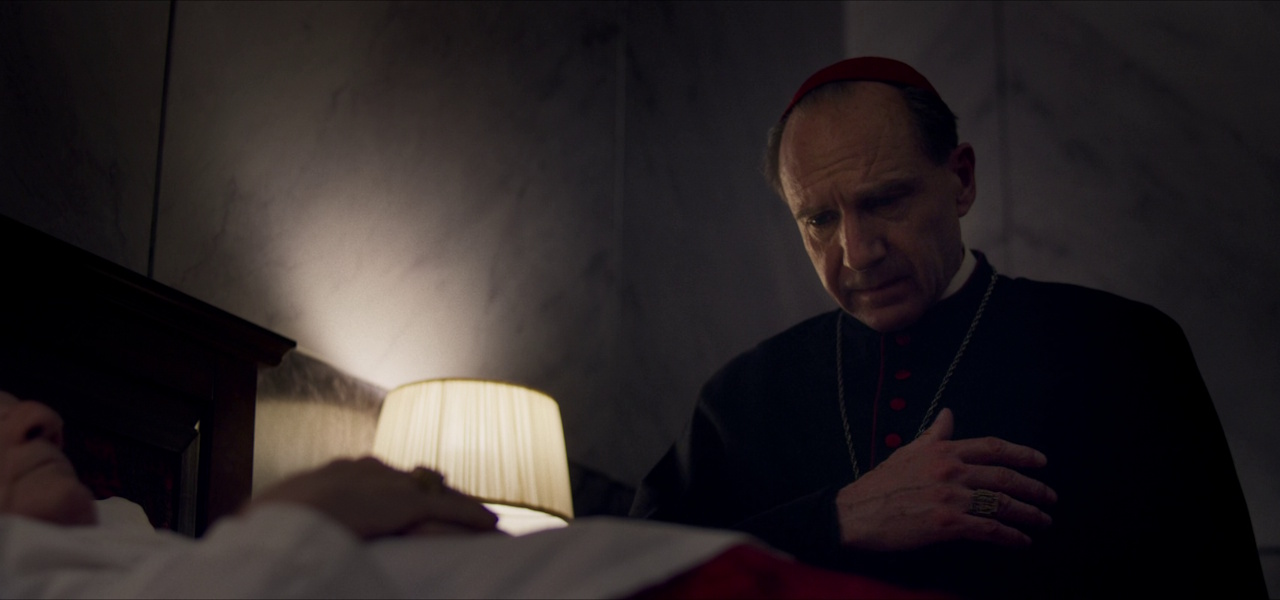 Still frame from Conclave (2024) showing Cardinal Lawrence praying over the body of the dead pope.