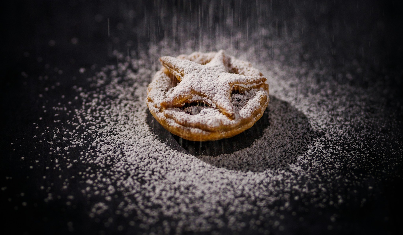 Stock photo of a mince pie on a dark slate being dusted with icing sugar.